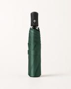 A folded dark green umbrella with a black handle, standing upright against a clean, off-white background.