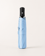 A light blue folded umbrella with a black handle, shown vertically against a clean, off-white background.